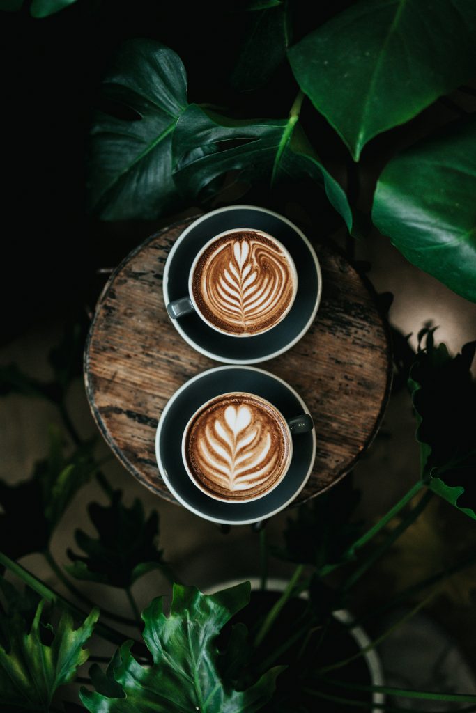 Das Bild zeigt zwei Tassen mit kunstvoll zubereitetem Latte Art, wobei auf der rechten oberen Ecke ein zartes Blattmuster zu sehen ist. Diese Darstellung symbolisiert die Kunstfertigkeit und Präzision, die in der Zubereitung eines perfekten Kaffees steckt. Es vermittelt den Fokus auf Qualität und Kreativität bei der Kaffeezubereitung und lädt die Betrachter ein, den besonderen Genuss von Kaffee auf eine visuelle Weise zu erleben.
