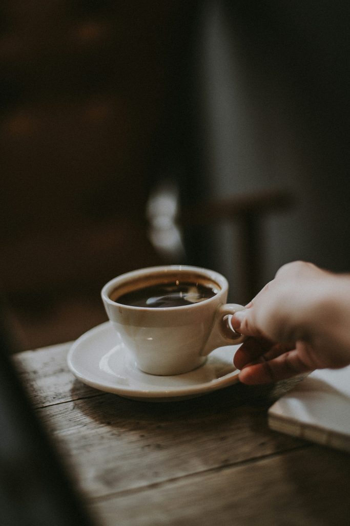 Das Bild zeigt eine Tasse schwarzen Kaffee in einem eleganten weißen Becher, die von einer sanften Frauenhand erreicht wird. Die Tasse steht in einer natürlichen, holzverkleideten Umgebung, die eine warme und gemütliche Atmosphäre schafft. Das Bild vermittelt ein Gefühl von Ruhe und Genuss, während es die Verbindung zwischen Mensch und Kaffee auf eine subtile, aber einladende Weise darstellt. Es symbolisiert den Moment der Entspannung und den bewussten Genuss einer Tasse Kaffee inmitten einer beruhigenden, natürlichen Umgebung.